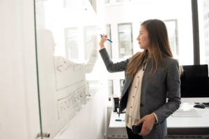 Femme en tenue professionnelle écrivant sur un tableau blanc dans un bureau lumineux, lors d’une séance de travail ou de coaching professionnel.