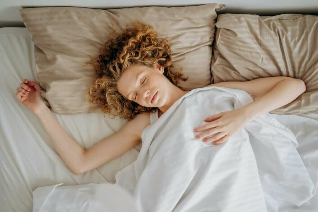 La sophrologie, une aide pour retrouver un sommeil de qualité Femme aux cheveux bouclés dormant d'un sommeil profond et réparateur, visage serein sur oreiller beige, symbole de qualité du sommeil grâce à la sophrologie : endormissement facile, cycles respectés, gestion réveils nocturnes.